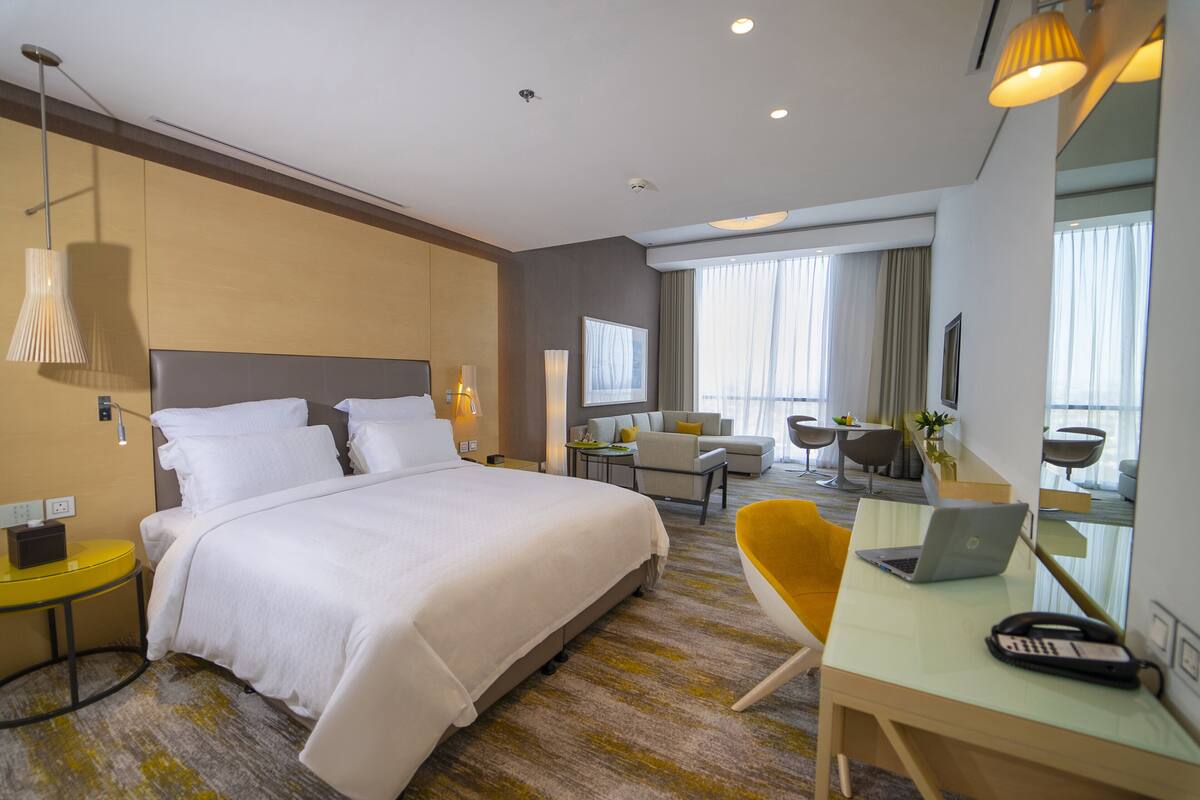 A hotel room with a large bed, a desk with a laptop, a chair, and a small table with a lamp.