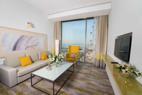 A modern hotel room with a sofa, armchair, coffee table, and a large window offering a city view. - Rotana Corniche (Dammam)