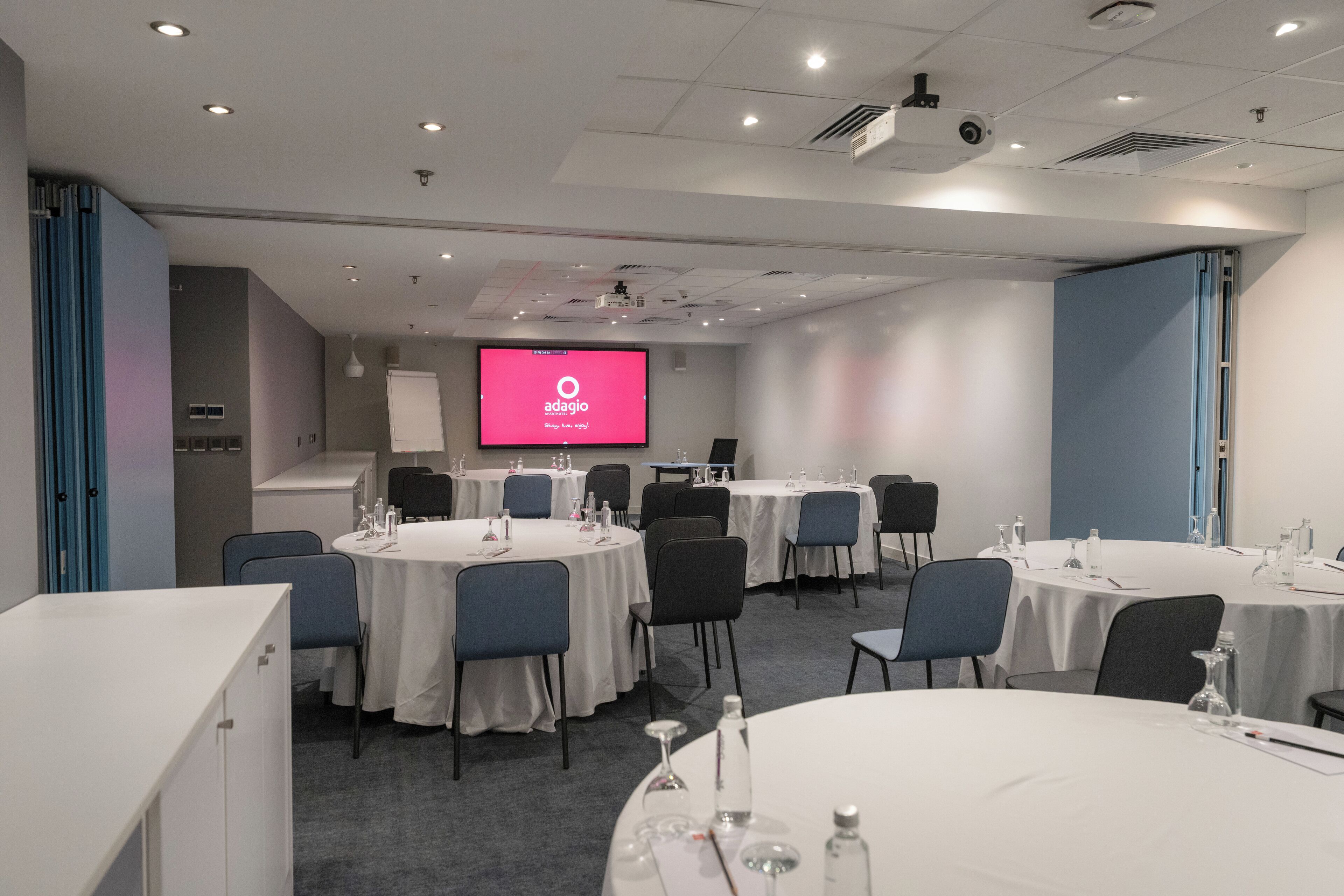 Un salón de conferencias con mesas redondas, sillas y una pantalla grande que muestra el logotipo de Adagio.