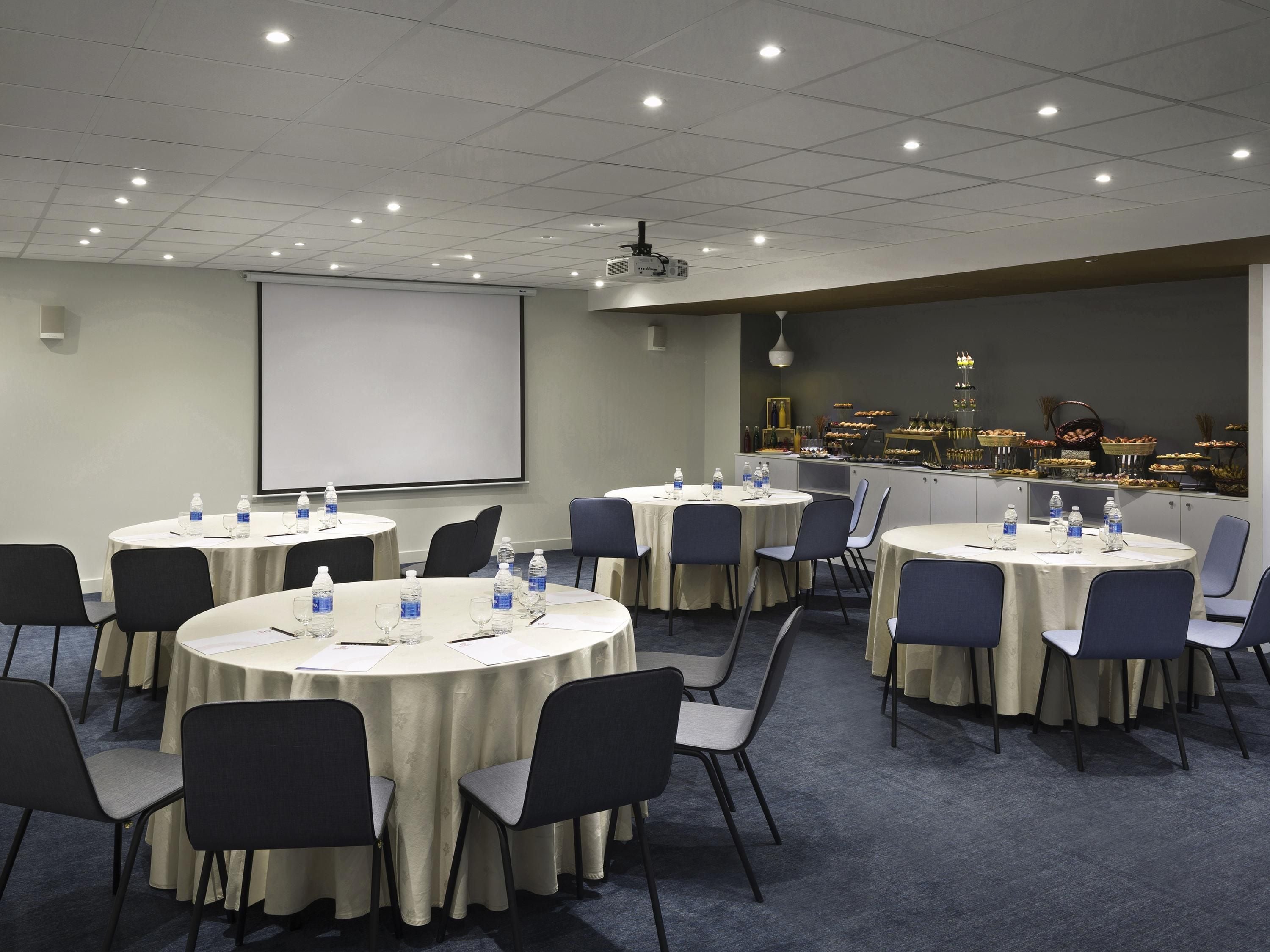 A conference room with multiple tables, chairs, a projector screen, and a buffet area with various food items.