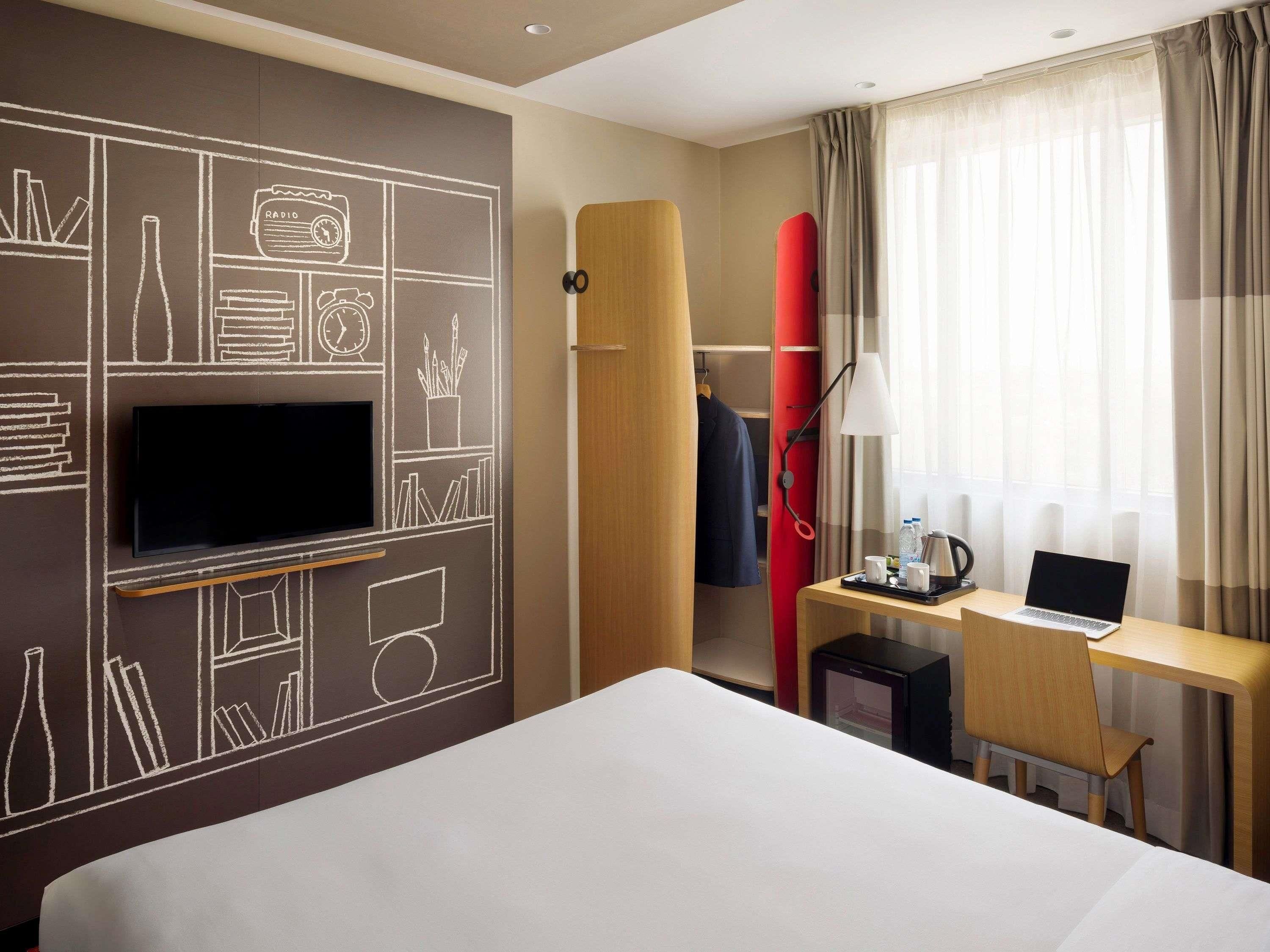Une chambre d’hôtel avec un lit, un bureau avec un ordinateur portable, une télévision, une planche de surf rouge et une armoire.
