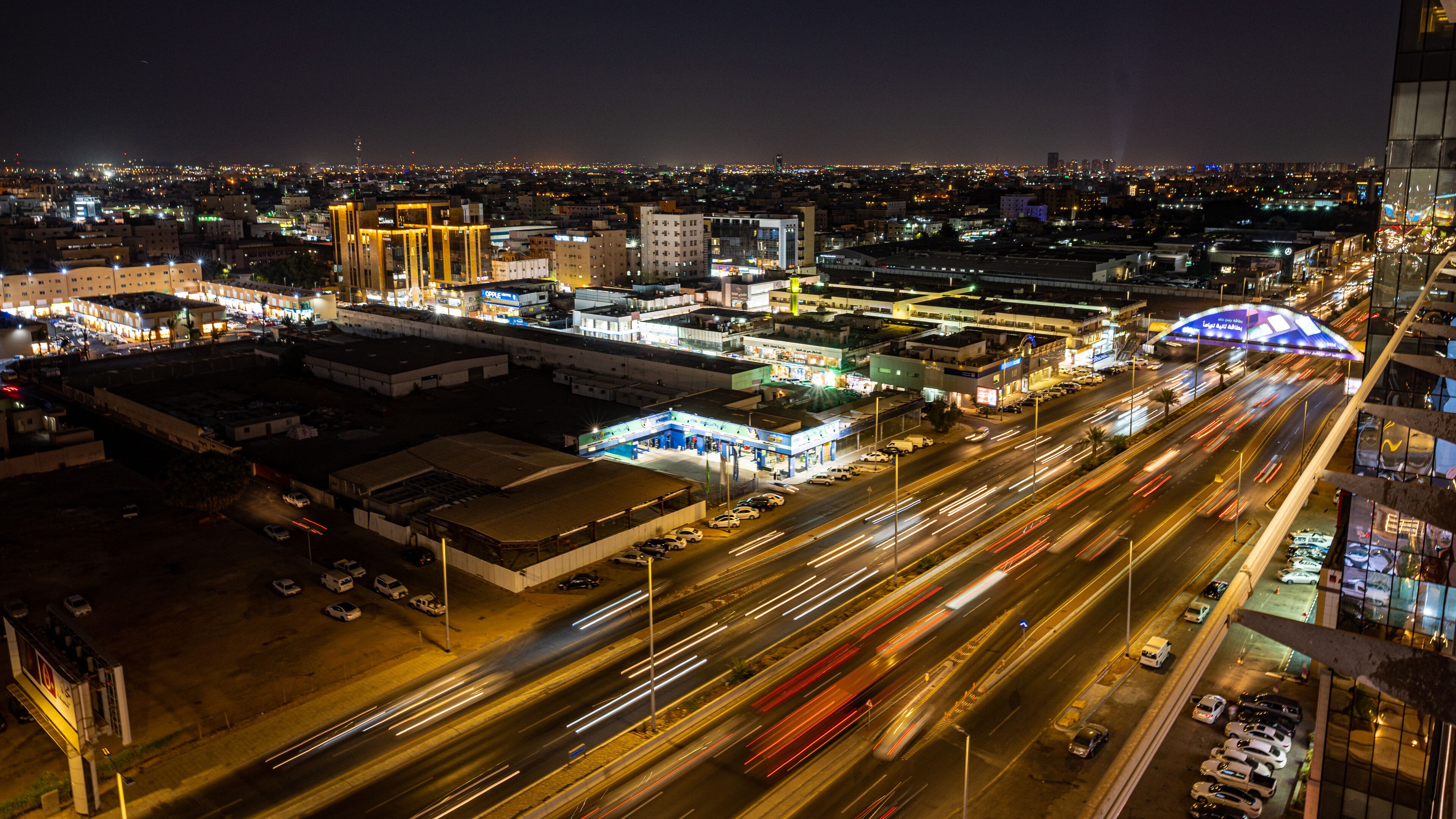 Panorama nocturno de una ciudad con edificios iluminados, una autopista con tráfico y un paso elevado para peatones.