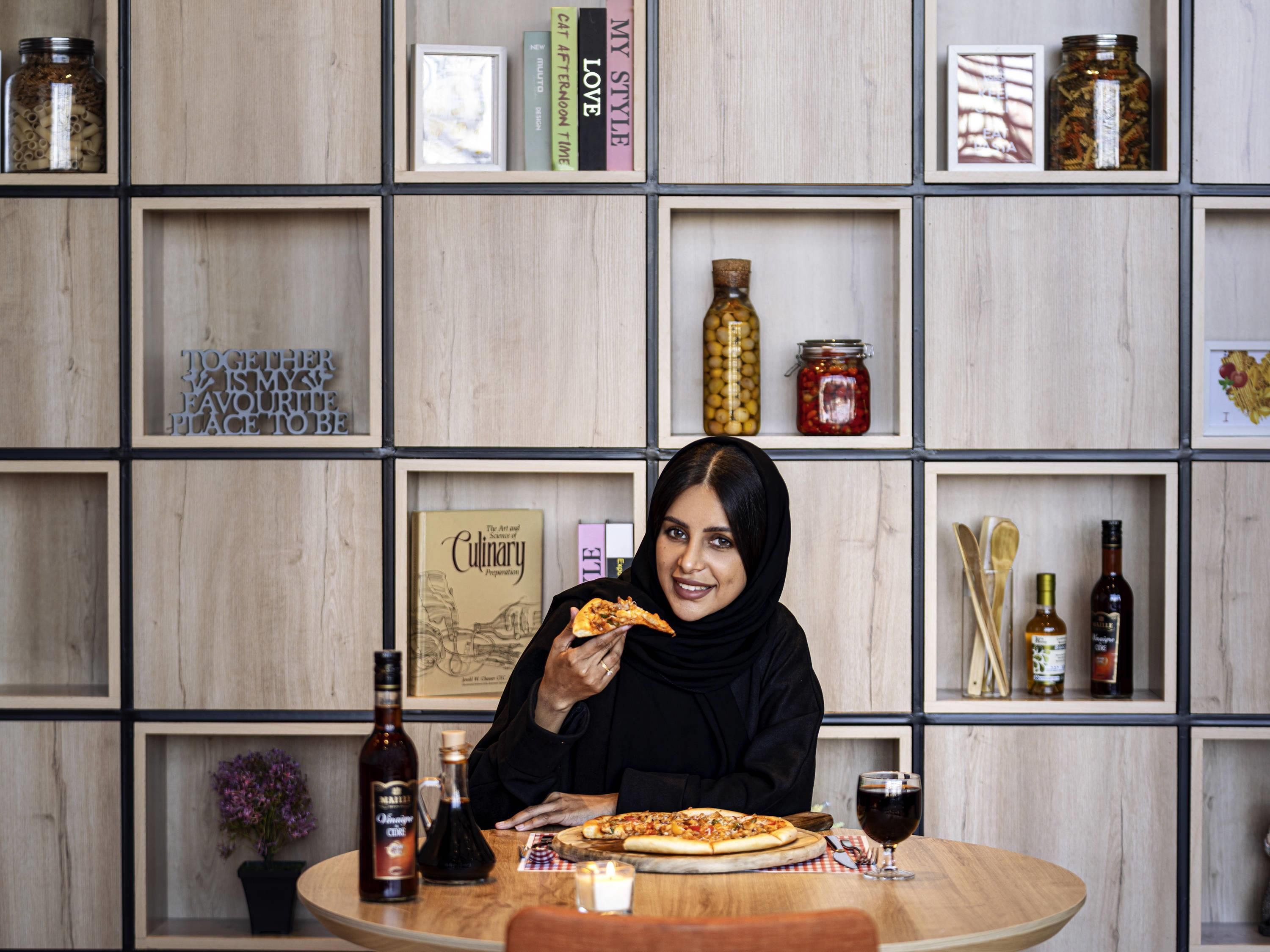 A person sitting at a wooden table with a pizza, a bottle of wine, and a candle. The background features a wooden bookshelf with various items.