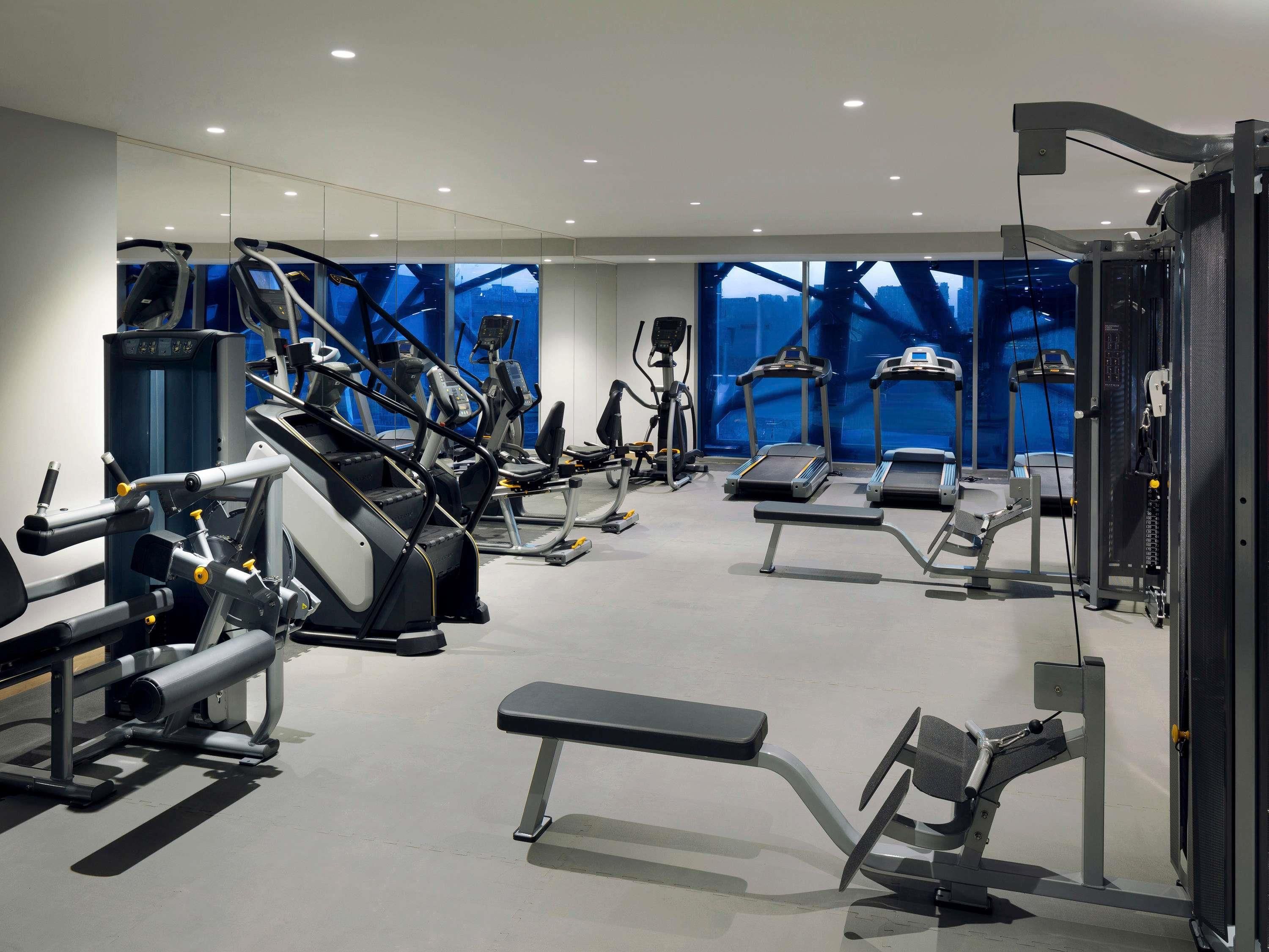 A modern gym with various exercise machines, a bench, and large windows with a city view.