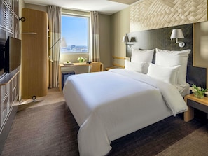 A hotel room with a large bed, a desk, a chair, a TV, and a city view.