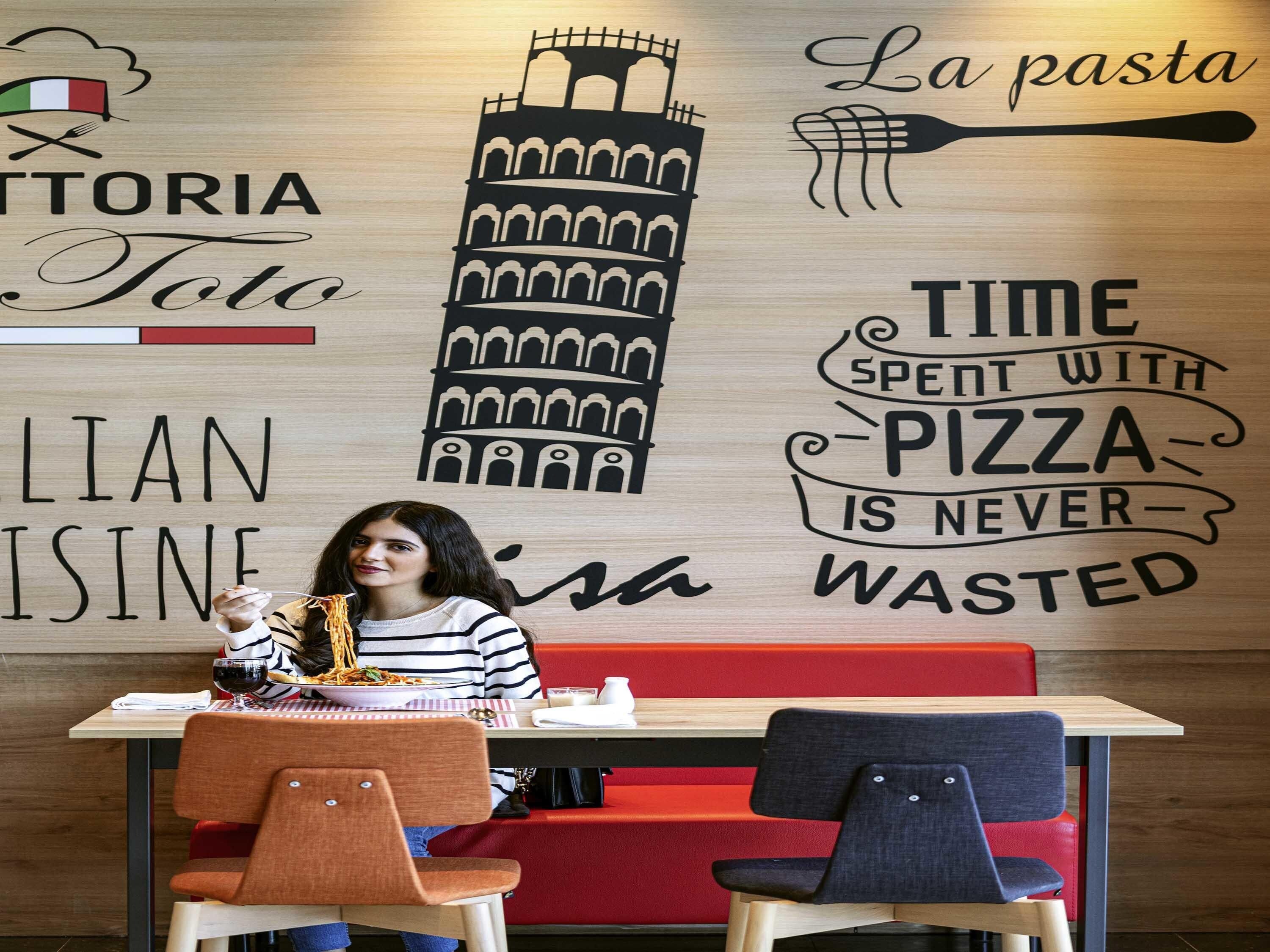 A restaurant interior with a wooden wall featuring Italian-themed graphics and text, a table with a person dining, and red and blue chairs.