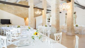 Un vaste hall doté de hauts plafonds et de colonnes en briques apparentes, aménagé avec des tables et des chaises blanches, et agrémenté de compositions florales.