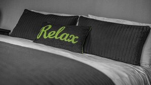 Una cama con colcha gris y dos almohadas, una con la palabra 'Relax' bordada en verde.