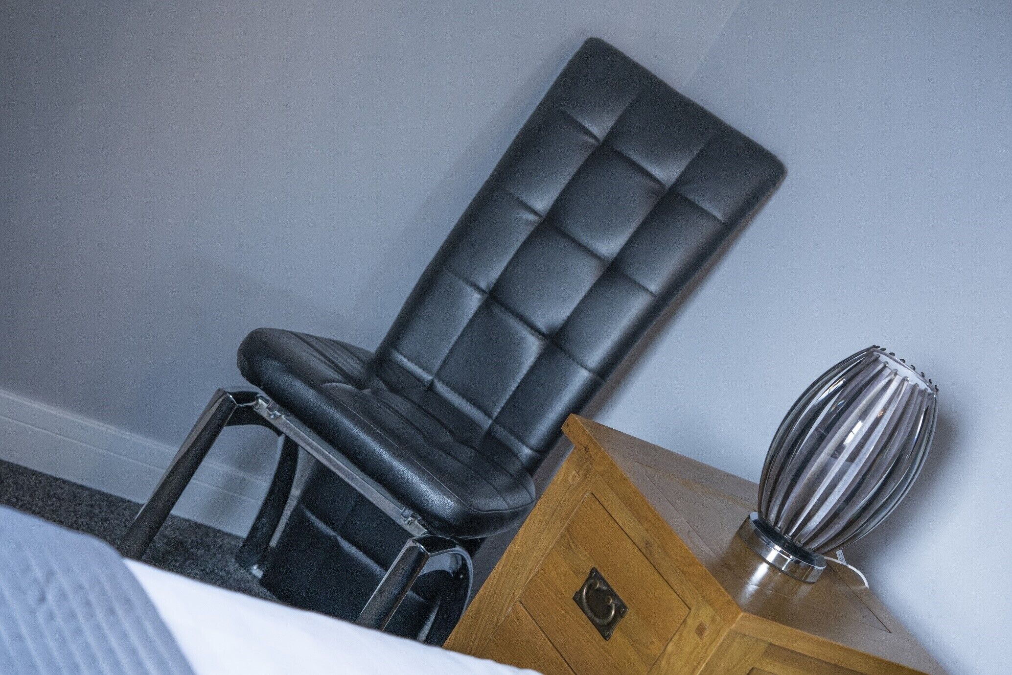 Un coin de pièce avec un fauteuil en cuir noir, une table de chevet en bois et un vase métallique.