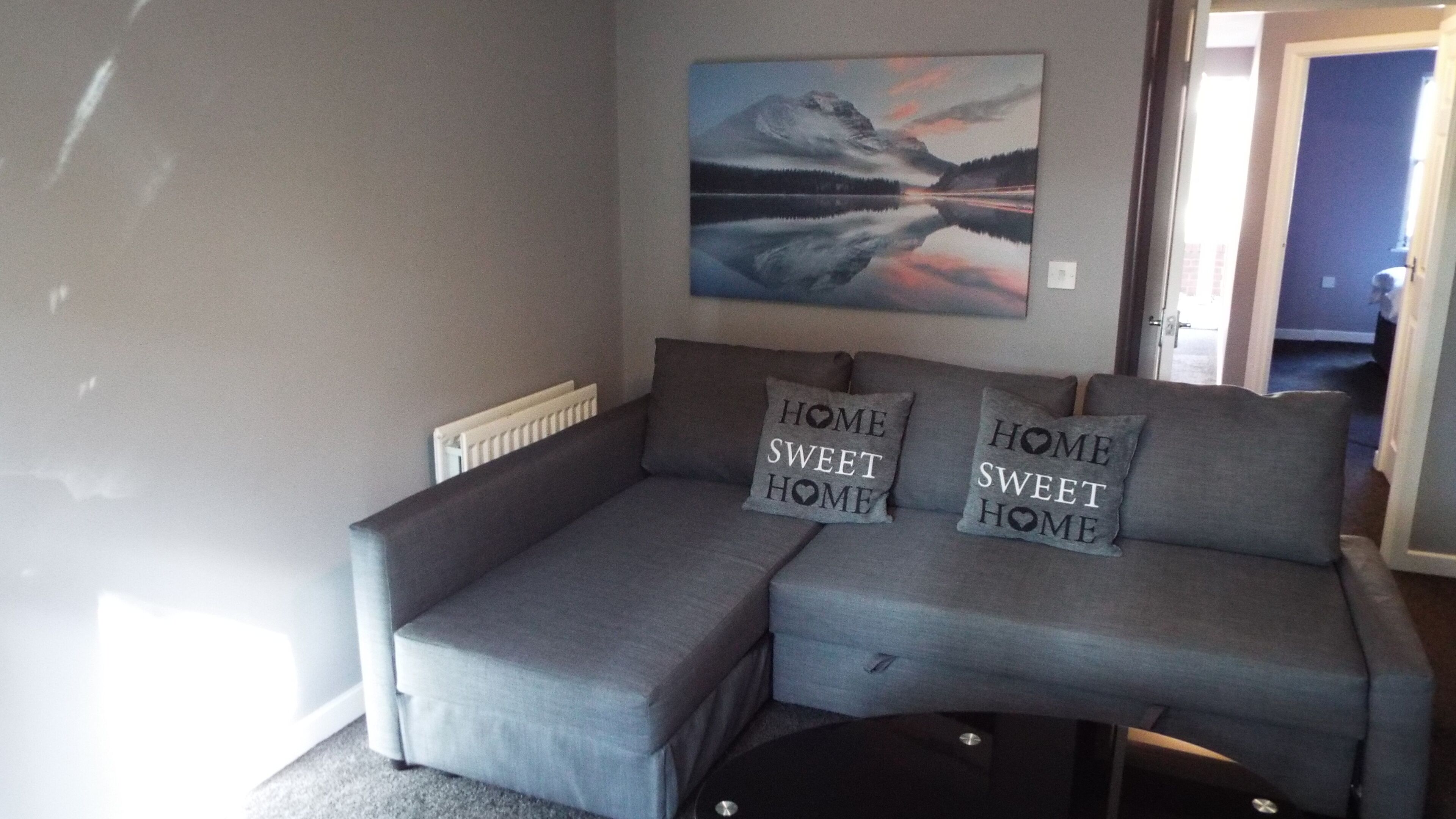 A grey sectional sofa with two pillows that read "HOME SWEET HOME" against a grey wall, with a painting above it.