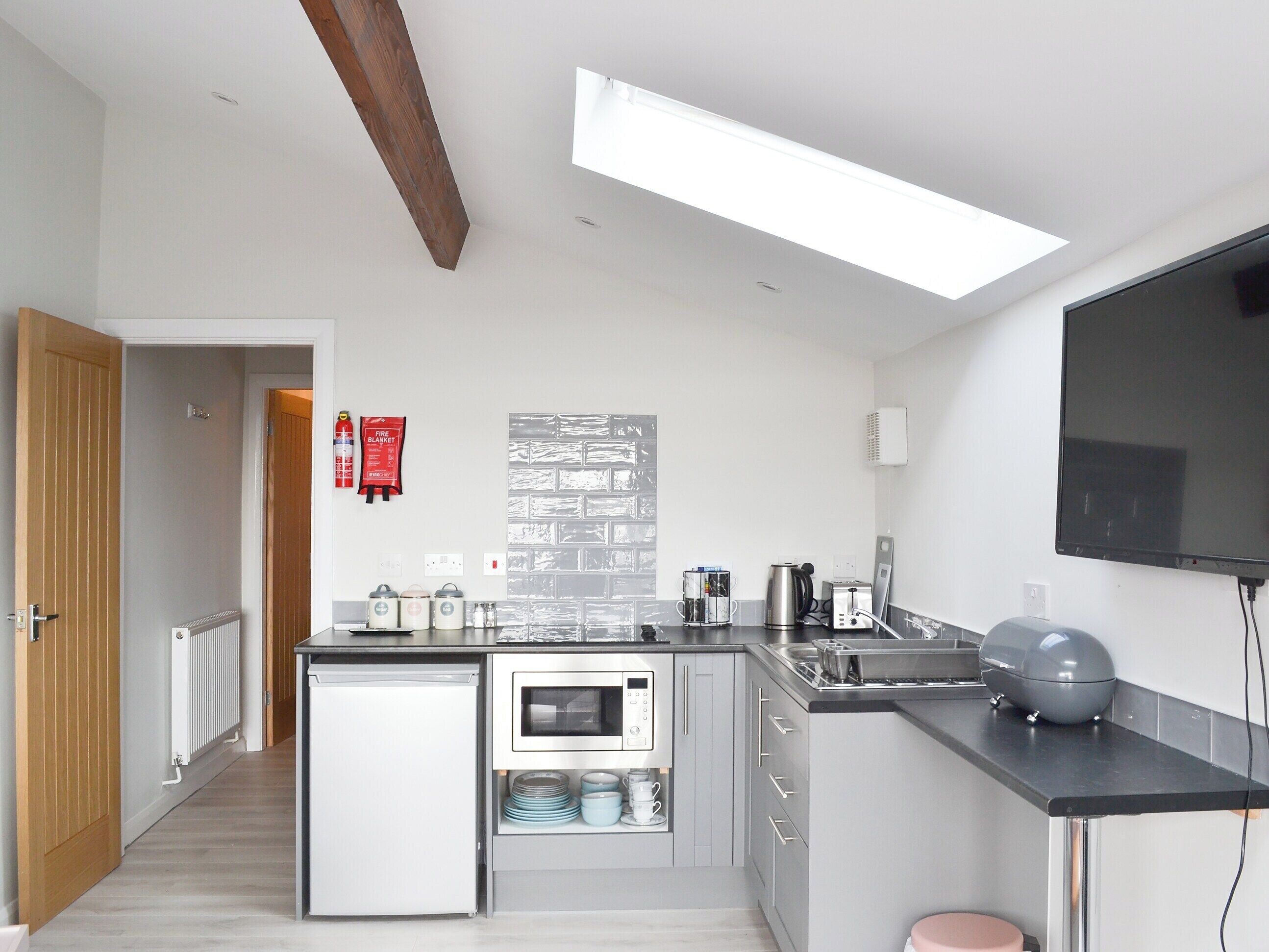 A modern kitchen with a built-in dishwasher, a microwave, and a coffee maker. There is a fire extinguisher on the wall and a flat-screen TV mounted above the counter.