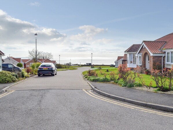A residential street with houses on both sides, a car parked on the left, and a clear view of the sea in the background.