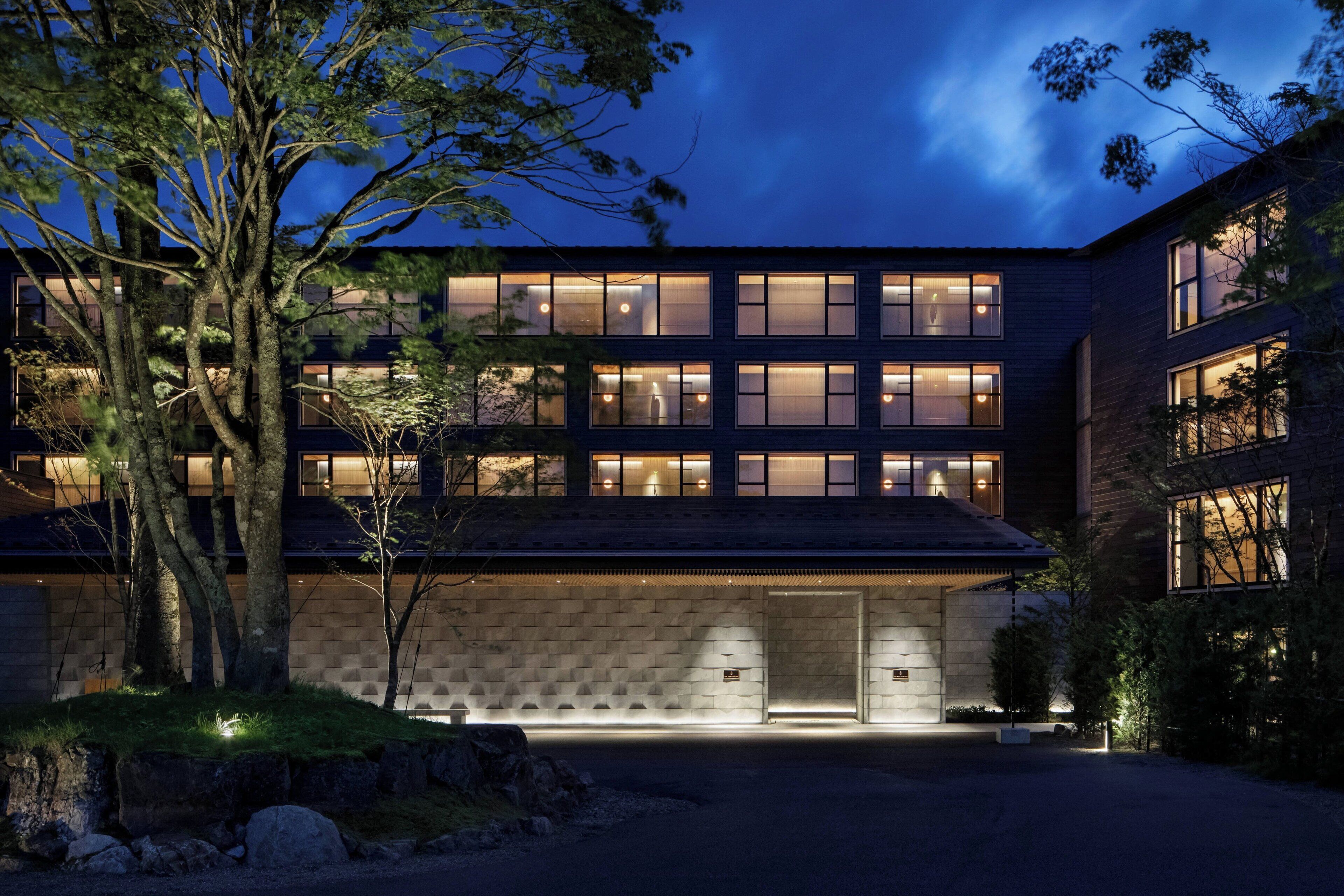 A modern building with multiple windows and a stone facade, surrounded by trees and a well-lit entrance.