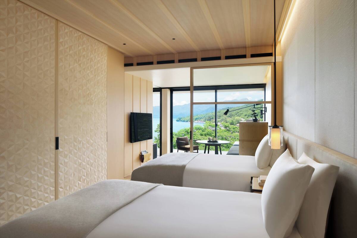 Habitación de hotel con dos camas, televisión, mesa de comedor con sillas y vistas a montañas a través de amplias ventanas.
