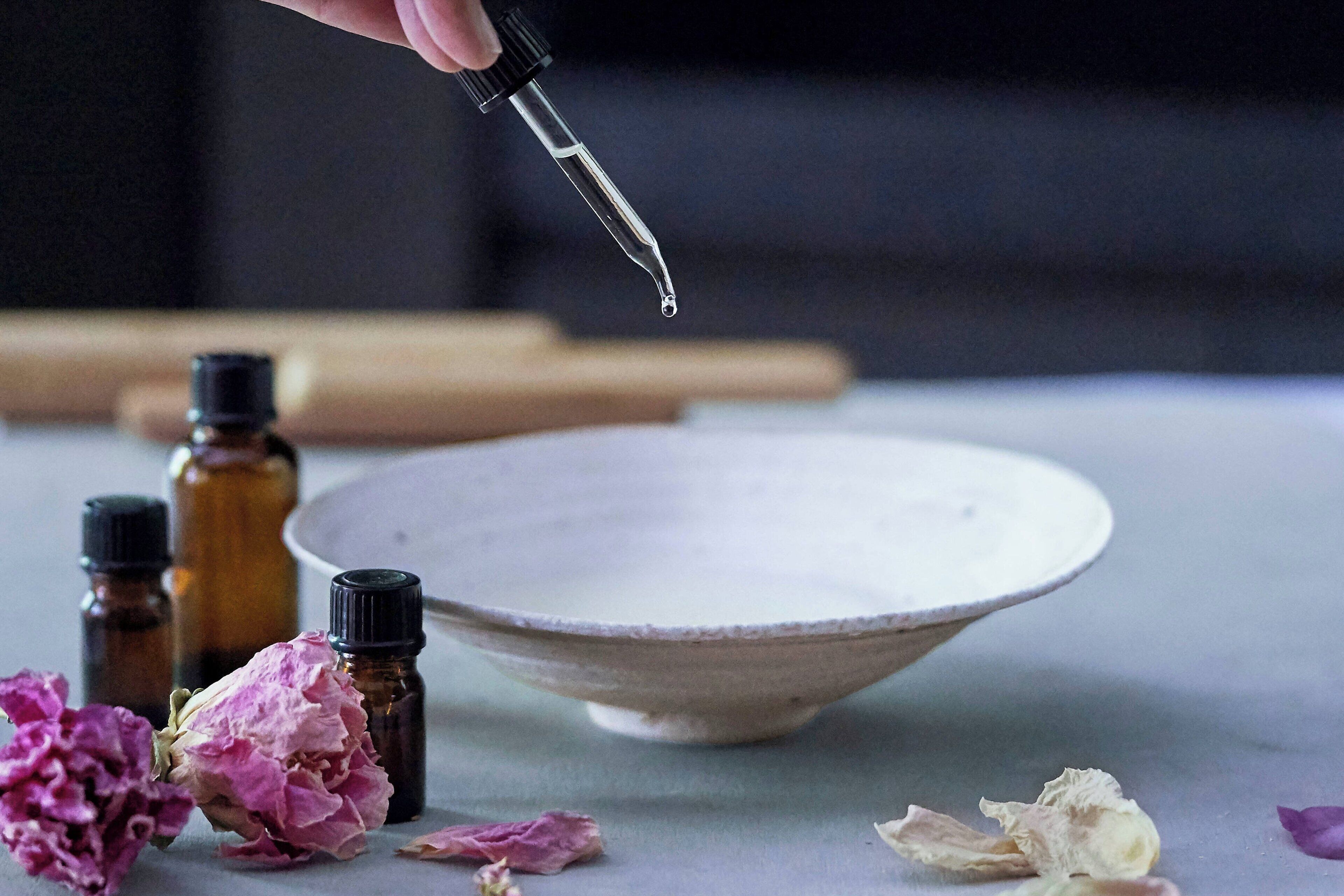 Un bol posé sur une table, avec un compte-gouttes et de petits flacons, qui pourraient contenir des huiles essentielles, et des fleurs séchées disposées autour.