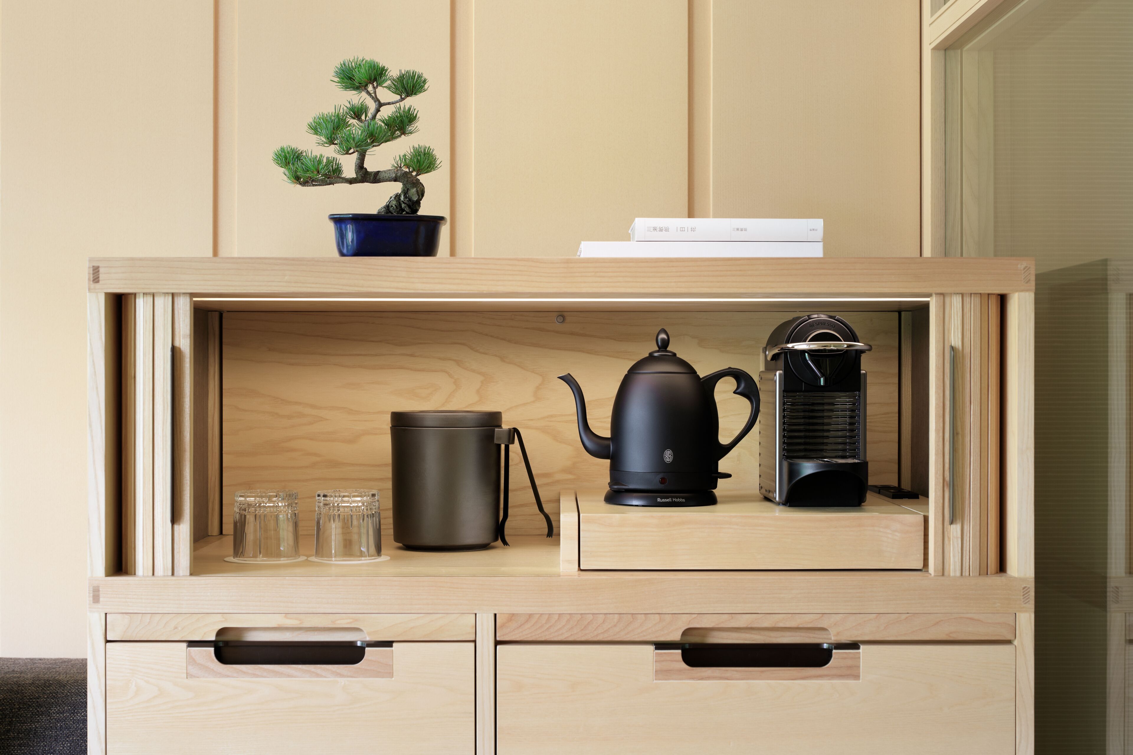 A wooden cabinet with a bonsai tree, a kettle, a coffee maker, and three glasses.
