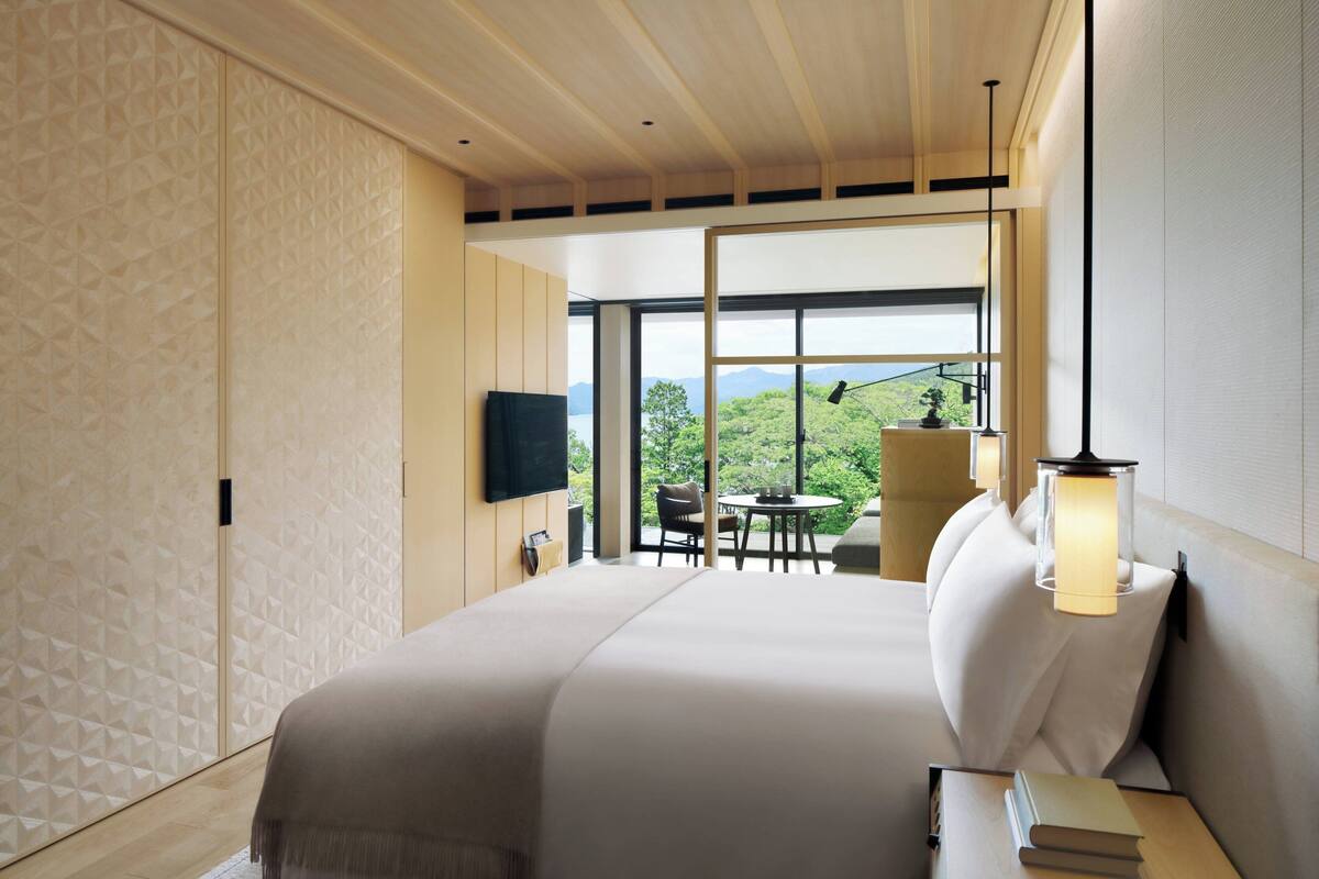 Una habitación de hotel moderna con una cama grande, un televisor montado en la pared, una zona de comedor con mesa y sillas, y vistas a zonas verdes a través de las ventanas.