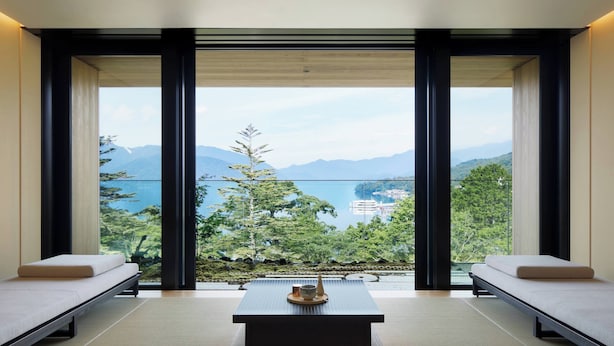 Habitación minimalista con suelo de tatami, una mesa baja con una taza, y vistas a montañas y agua.
