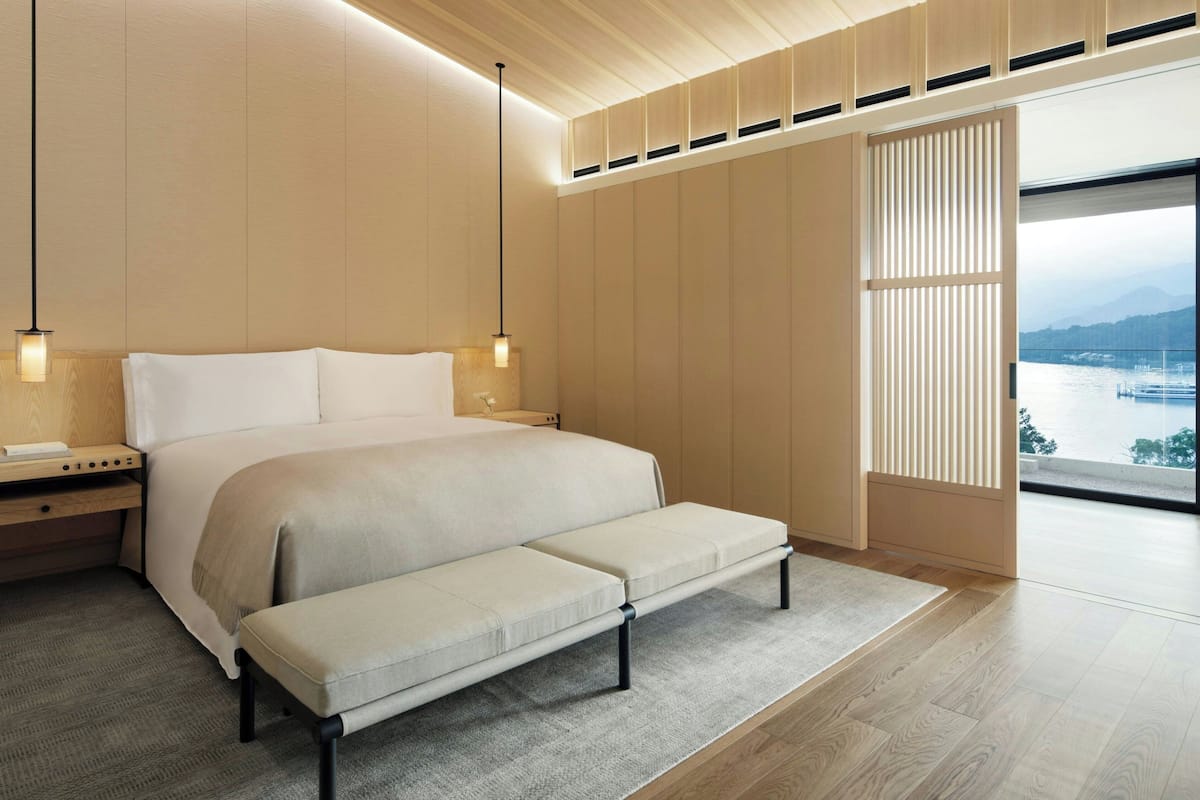 Un dormitorio moderno con una cama grande, mesitas de noche, un banco, vista a una masa de agua a través de grandes ventanales y un techo de madera.