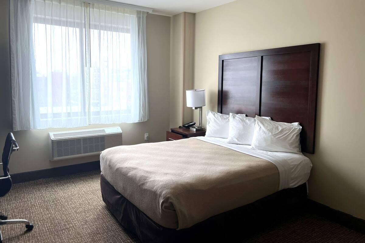 A hotel room with a large bed, a bedside lamp, a window with sheer curtains, and a wall-mounted air conditioner.
