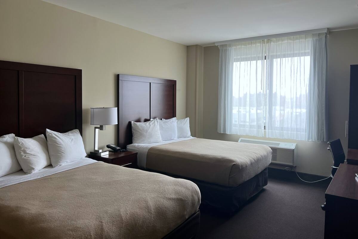 A hotel room with two beds, a desk, a chair, a lamp, and a window with sheer curtains.