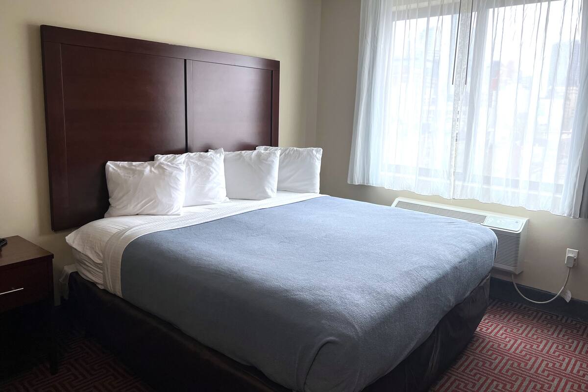 A hotel room with a large bed, a wooden headboard, white pillows, a grey bedspread, a window with sheer curtains, and a radiator.