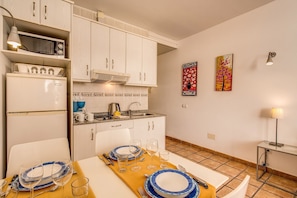 A compact kitchen with white cabinets, a microwave, and a sink. There are two wall-mounted paintings and a table set for dining.