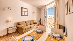 A modern living room with a sofa, a dining table set for two, and a large window with a view of buildings. - GRAN CANARIA HOME (7), ARGUINEGUIN BEACH AND SUN (Arguineguin)