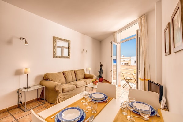 A modern living room with a sofa, a dining table set for two, and a large window with a view of buildings.
