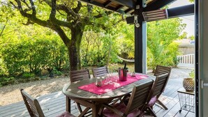 Une terrasse extérieure avec une table et des chaises en bois, entourée d’une végétation luxuriante et d’un grand arbre.