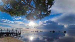 Une plage paisible avec une jetée en bois qui s’avance dans l’eau, des bateaux ancrés à proximité, et un ciel dégagé où la lumière du soleil se reflète sur l’eau.