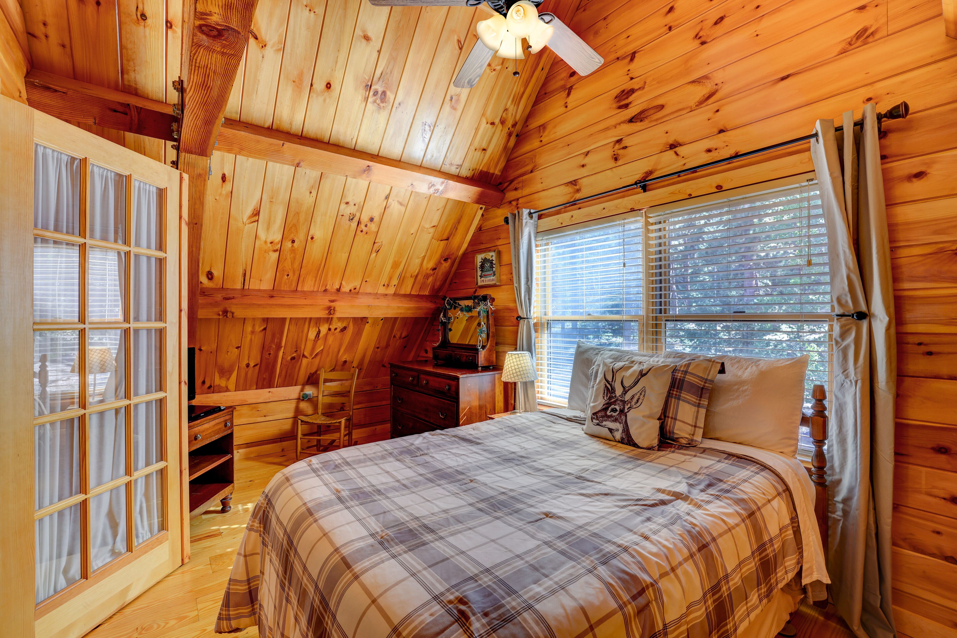 A cozy cabin bedroom with a plaid bedspread, wooden walls, a ceiling fan, and a window with blinds.