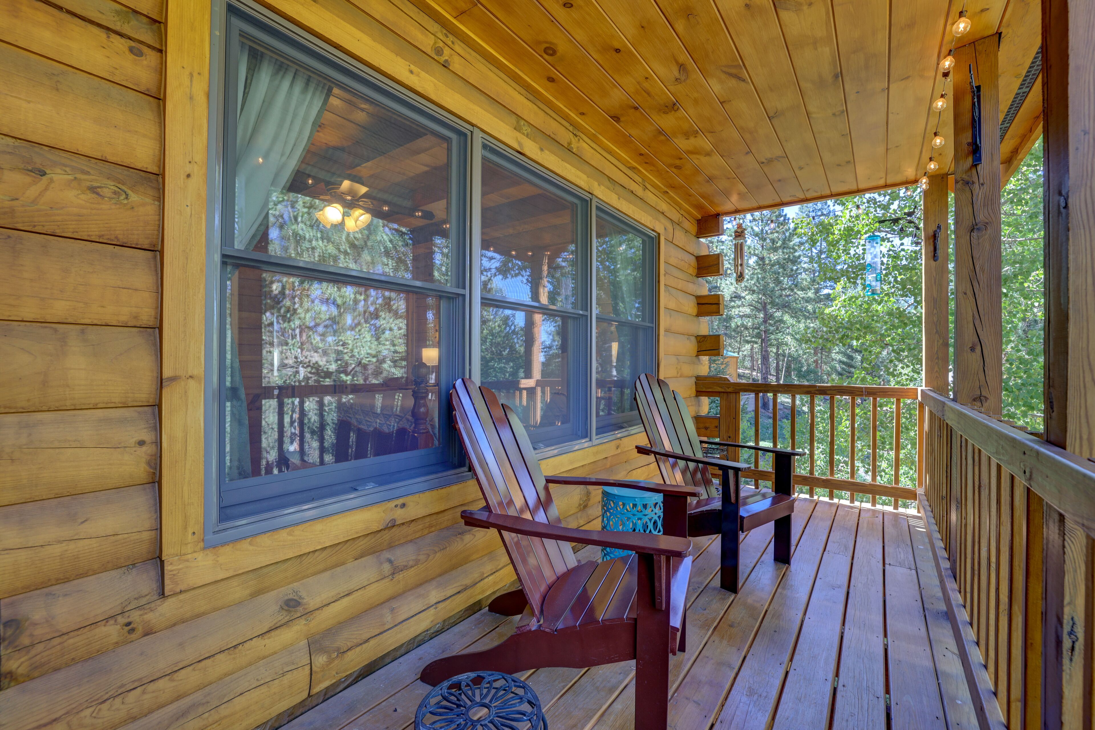 A wooden deck with two Adirondack chairs and a table, large windows, and a view of trees.