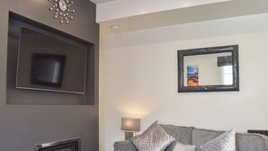 A modern living room with a grey sofa, a flat-screen TV mounted on the wall, a fireplace, and a mirror reflecting a cityscape.