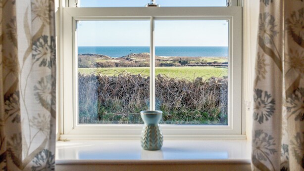 A window with a view of a coastal landscape, a vase on the windowsill, and patterned curtains.