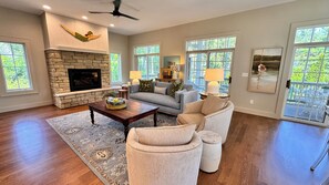 A spacious living room with a stone fireplace, a sofa set, a coffee table, and a large window with a view of greenery.