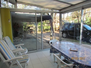 A covered patio with a table and chairs, a car parked outside, and a view of a garden and trees.