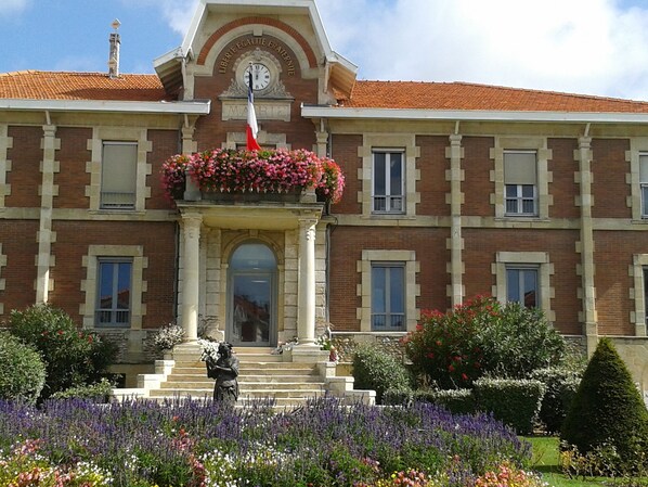 A building with a clock, a statue, and a flower bed in front. - ATLANTIC VILLA (Soulac-sur-Mer)
