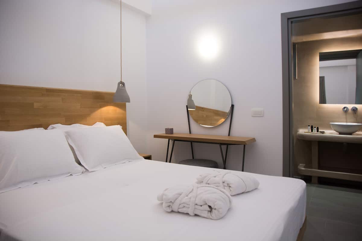 A hotel room with a neatly made bed, a bedside table with a lamp, a round mirror, and a bathroom with a sink and a large bowl.