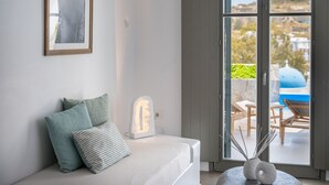 A modern bedroom with a bed, pillows, a bedside lamp, and a view of an outdoor area with a barbecue and seating. - Amphitrite Suites Santorini (Santorini)