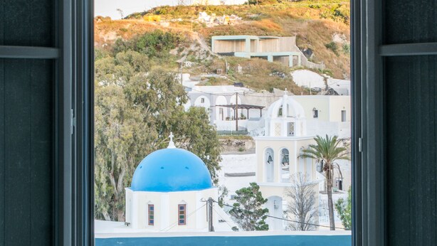 A view through a window of a building with a blue dome, a pool, and a hillside with buildings. - Amphitrite Suites Santorini (Santorini)