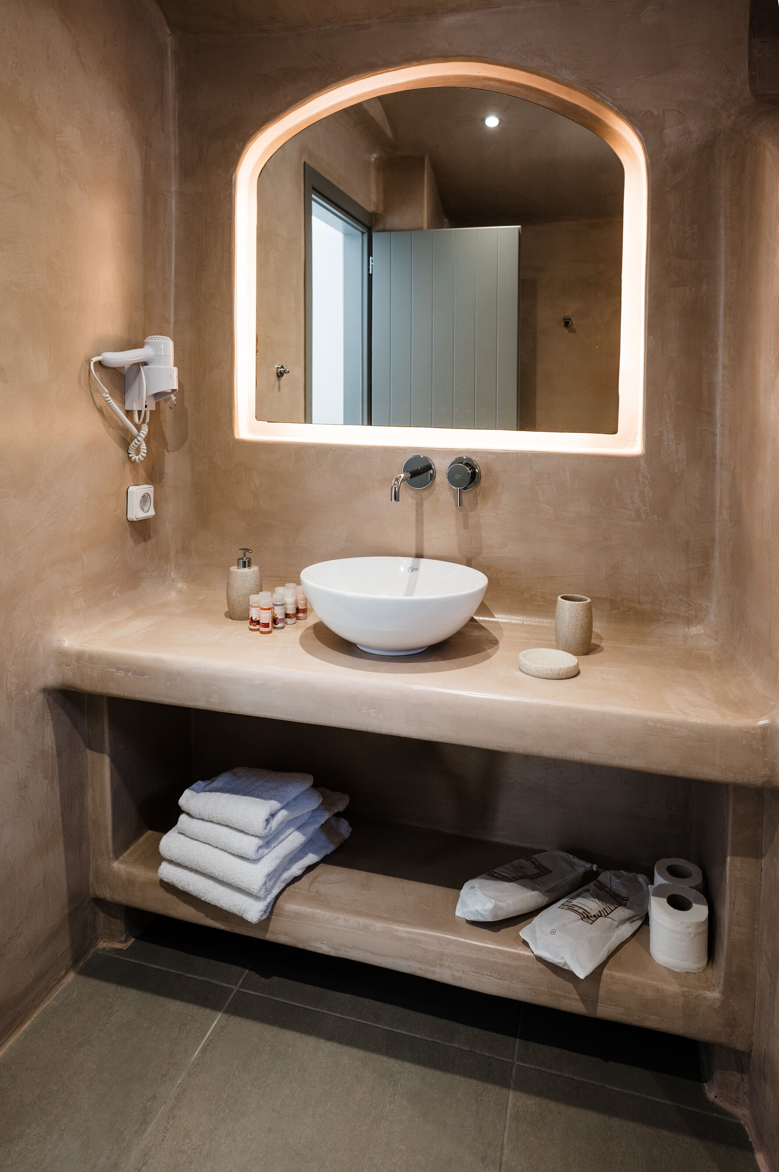 Een badkamer met een groot, rond wastafel, een spiegel met een boogvormig frame, en een plank onder de wastafel met handdoeken en toiletartikelen.