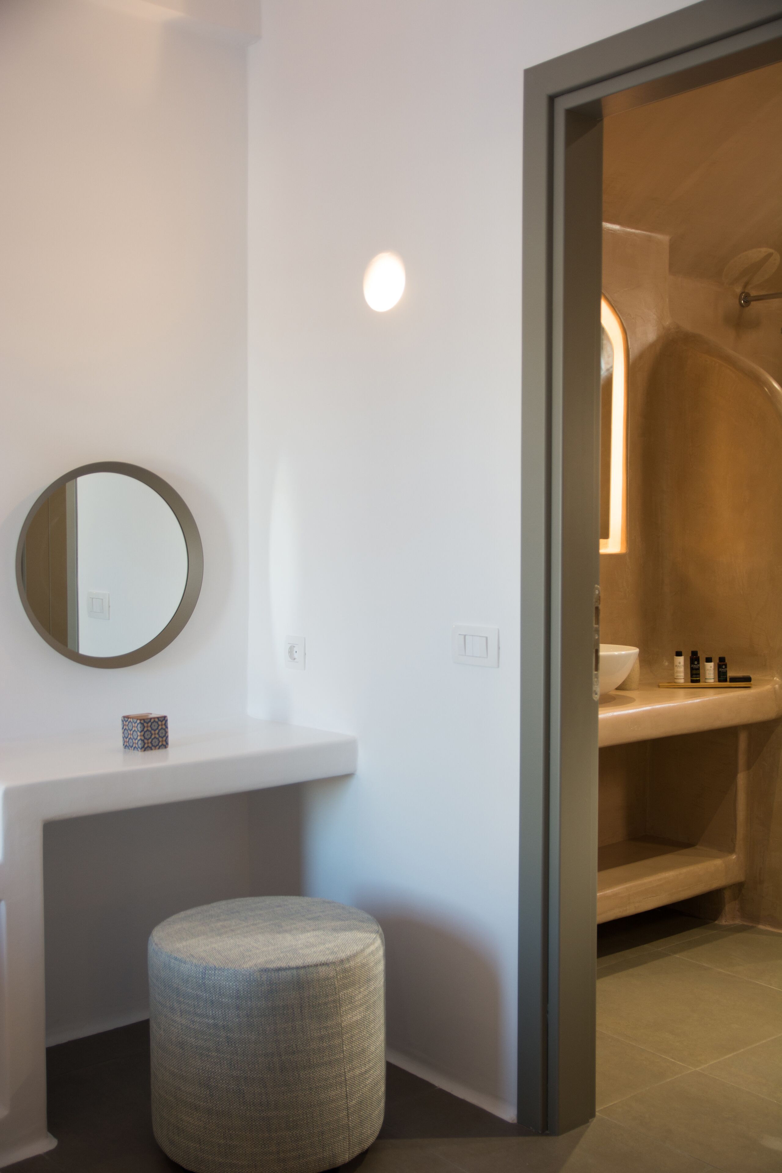 Une salle de bain moderne avec un miroir rond, un petit tabouret et une commode où sont rangés les articles de toilette.