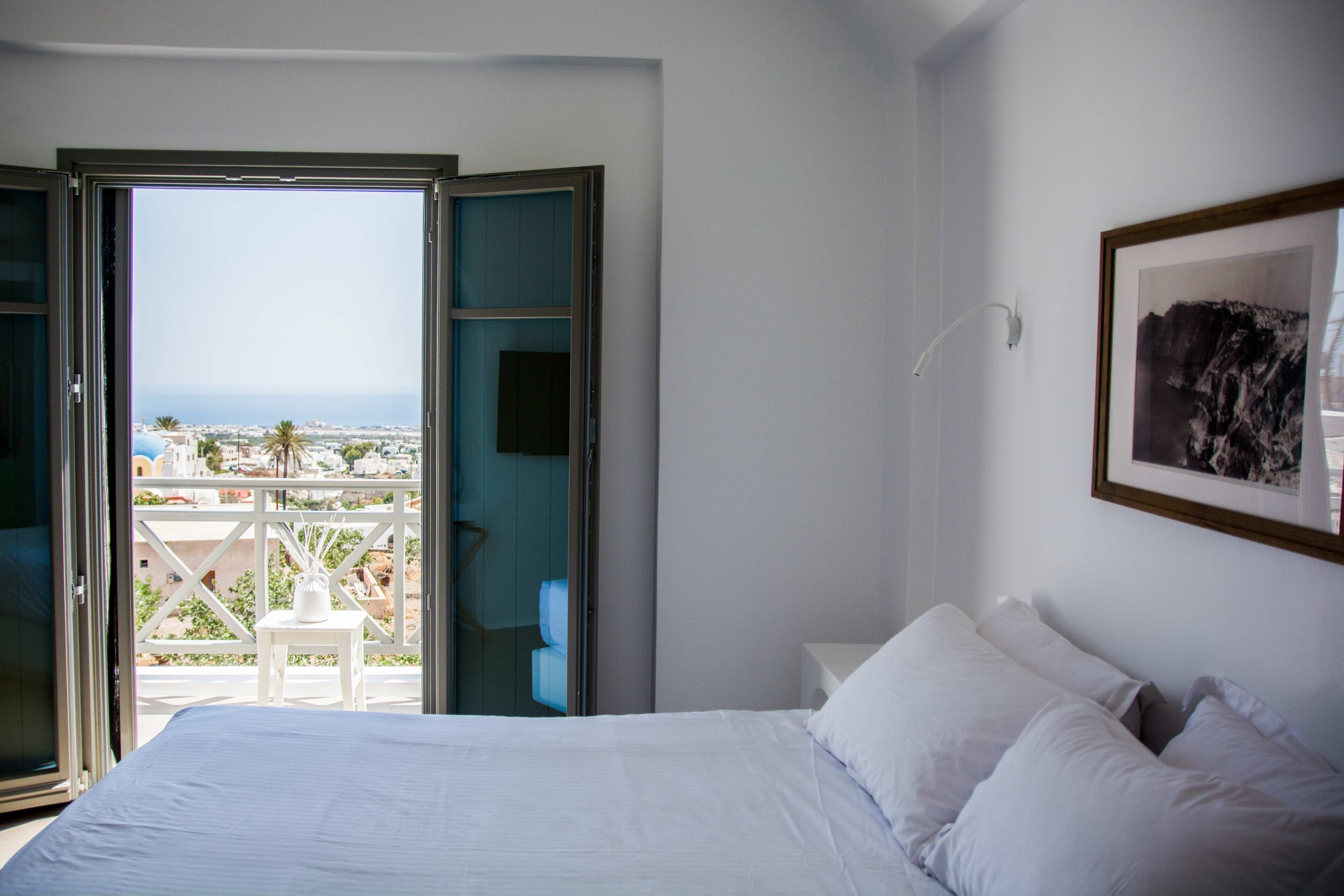 A bedroom with a bed, a nightstand, a framed picture on the wall, and a balcony with a view of the city and ocean.