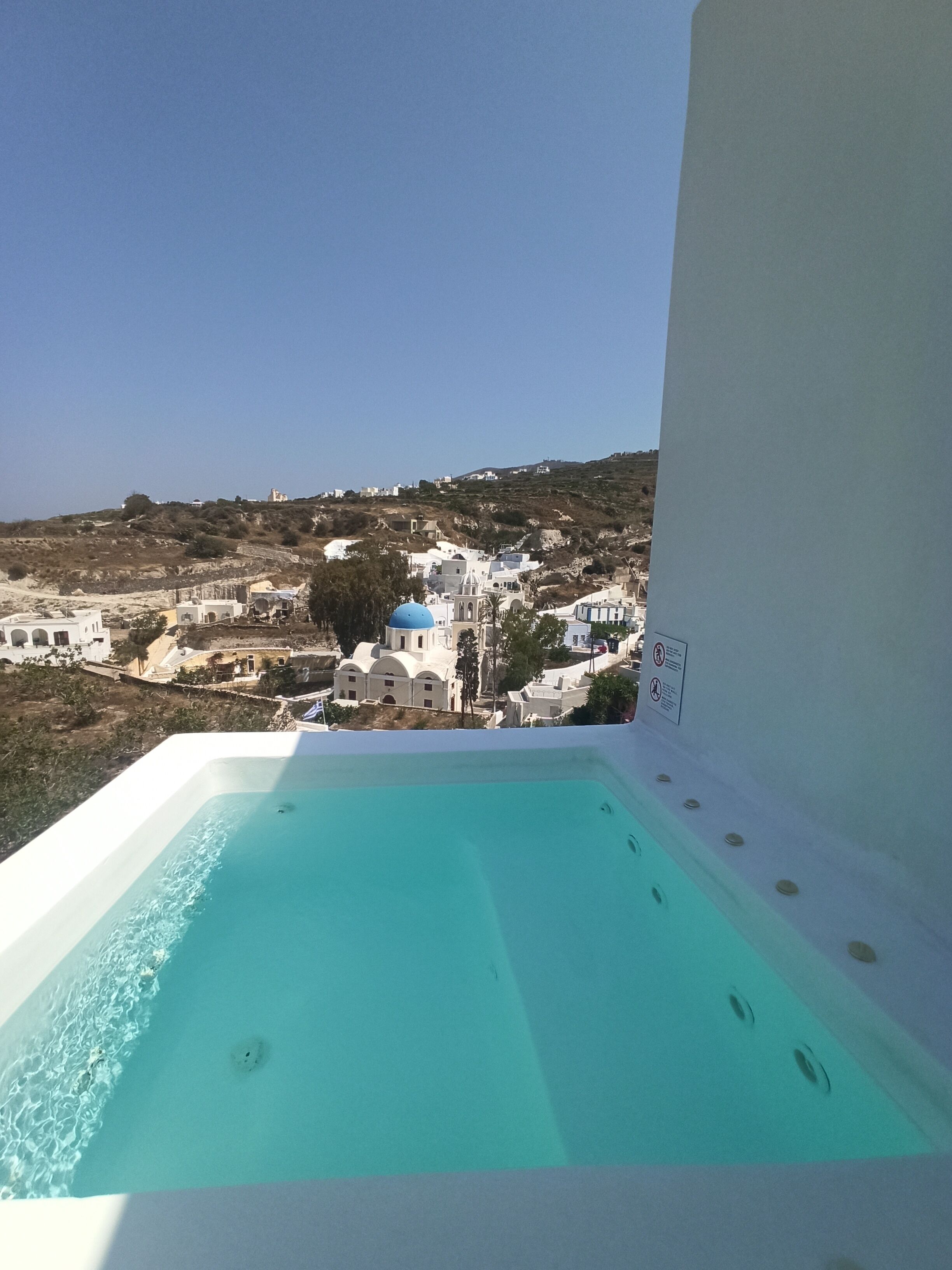 Piscina azul límpida com vista para uma vila costeira, incluindo edifícios brancos e uma igreja com cúpula azul.