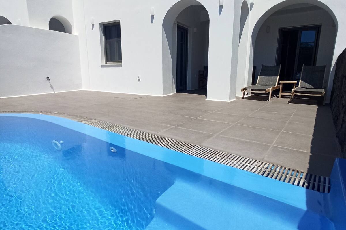 A swimming pool with clear blue water, a white building with arched doorways, and a seating area with chairs.