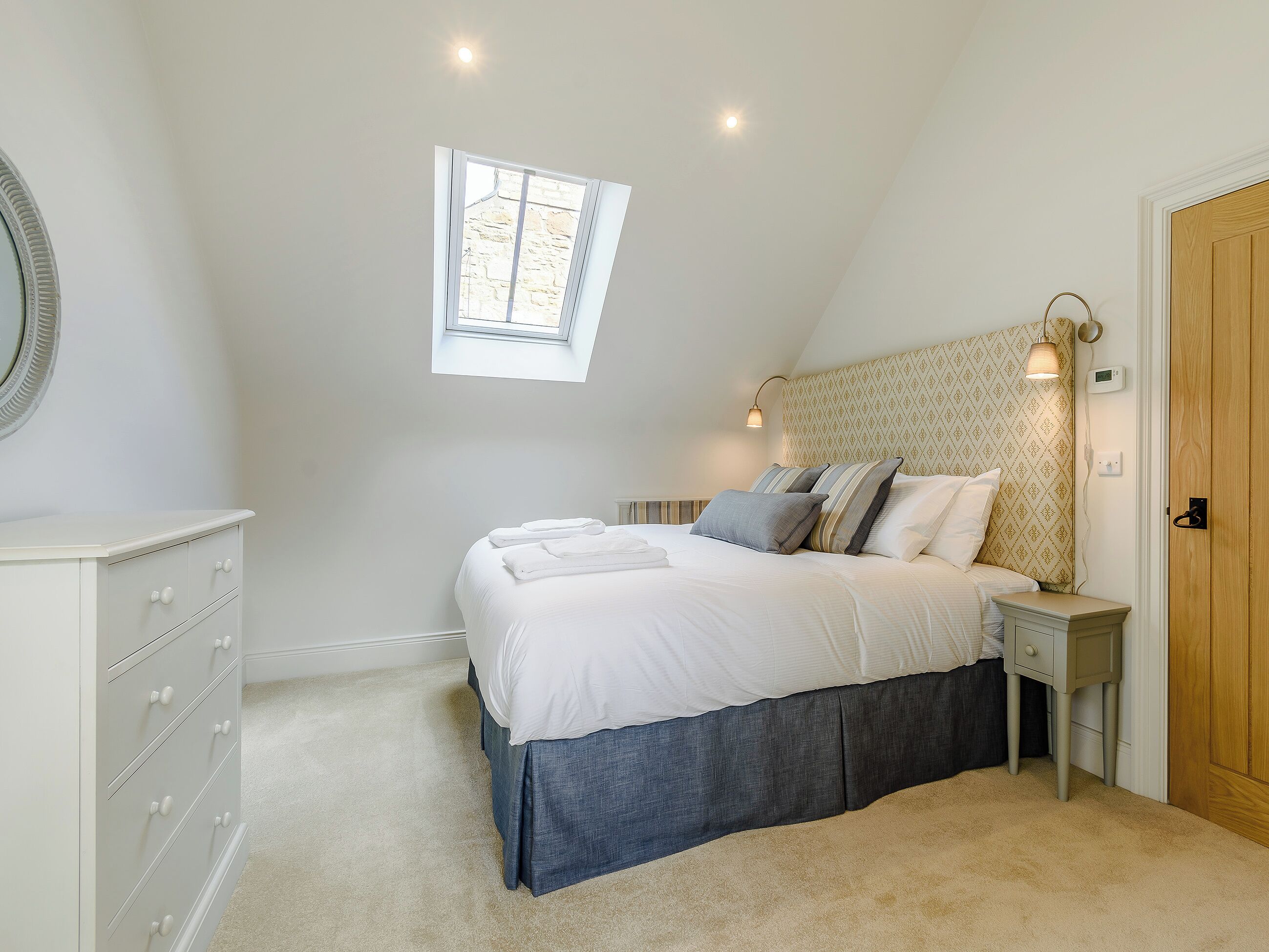 Un dormitorio con cama, cómoda, mesita de noche, lámpara de pared y ventana pequeña.