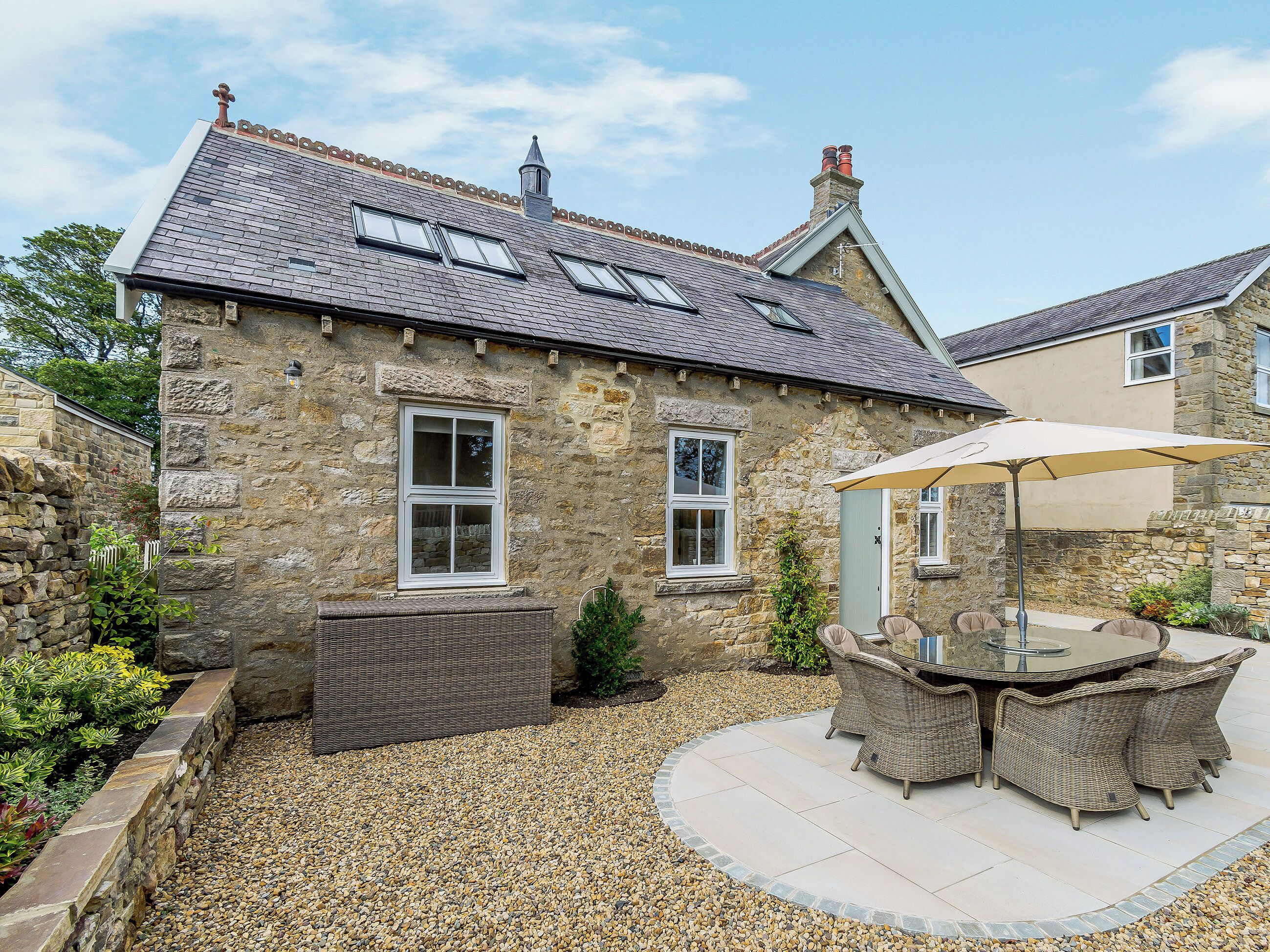 A stone building with a tiled roof, a patio with wicker furniture, and a gravel ground.