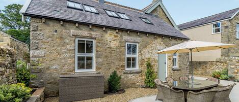 A stone building with a tiled roof, a patio with wicker furniture, and a gravel ground.