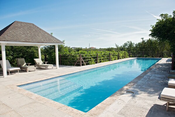 Une piscine rectangulaire avec un gazebo blanc et des chaises longues d’un côté.